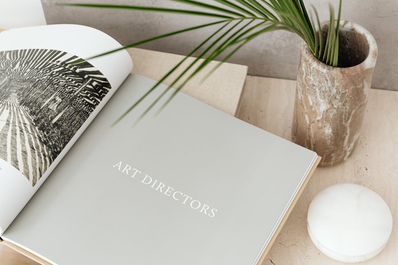 services-01 Open art book beside a decorative marble vase and palm leaf, showcasing modern aesthetics.